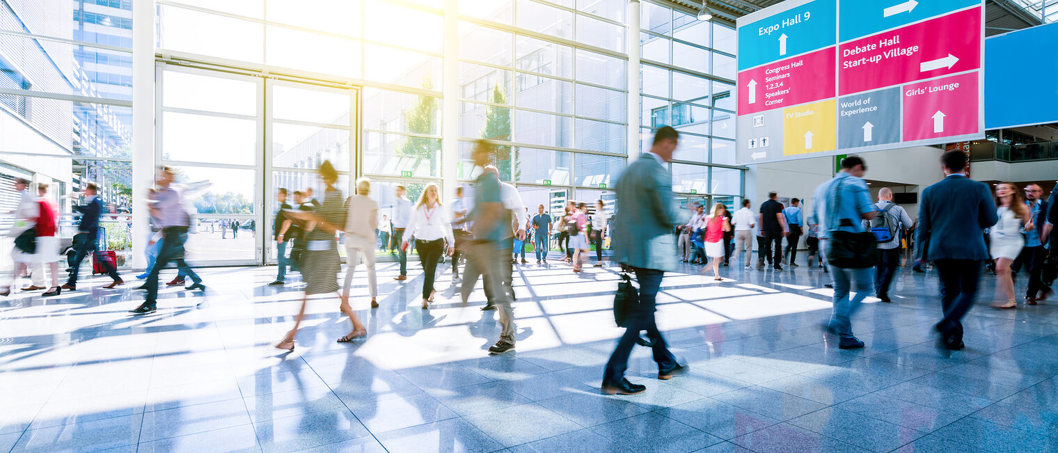 Menschenmenge in einem hellen Messe- oder Kongresszentrum, im Hintergrund große Fensterfront und farbige Wegweiser.