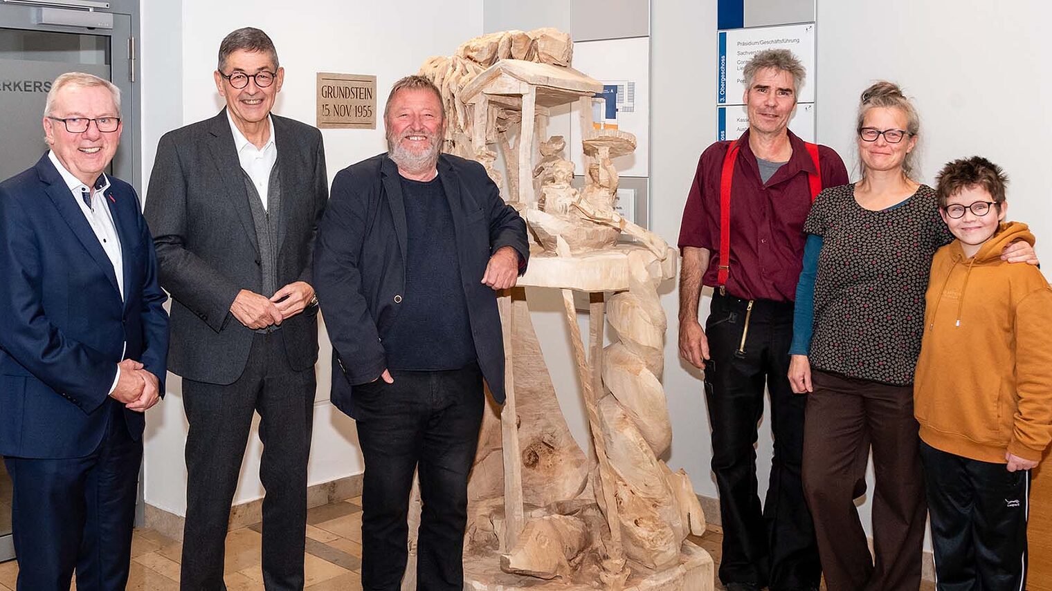 Handwerkskammervertreter und Künstler stehen neben einer Skulptur die aus Holz geschnitzt ist. 