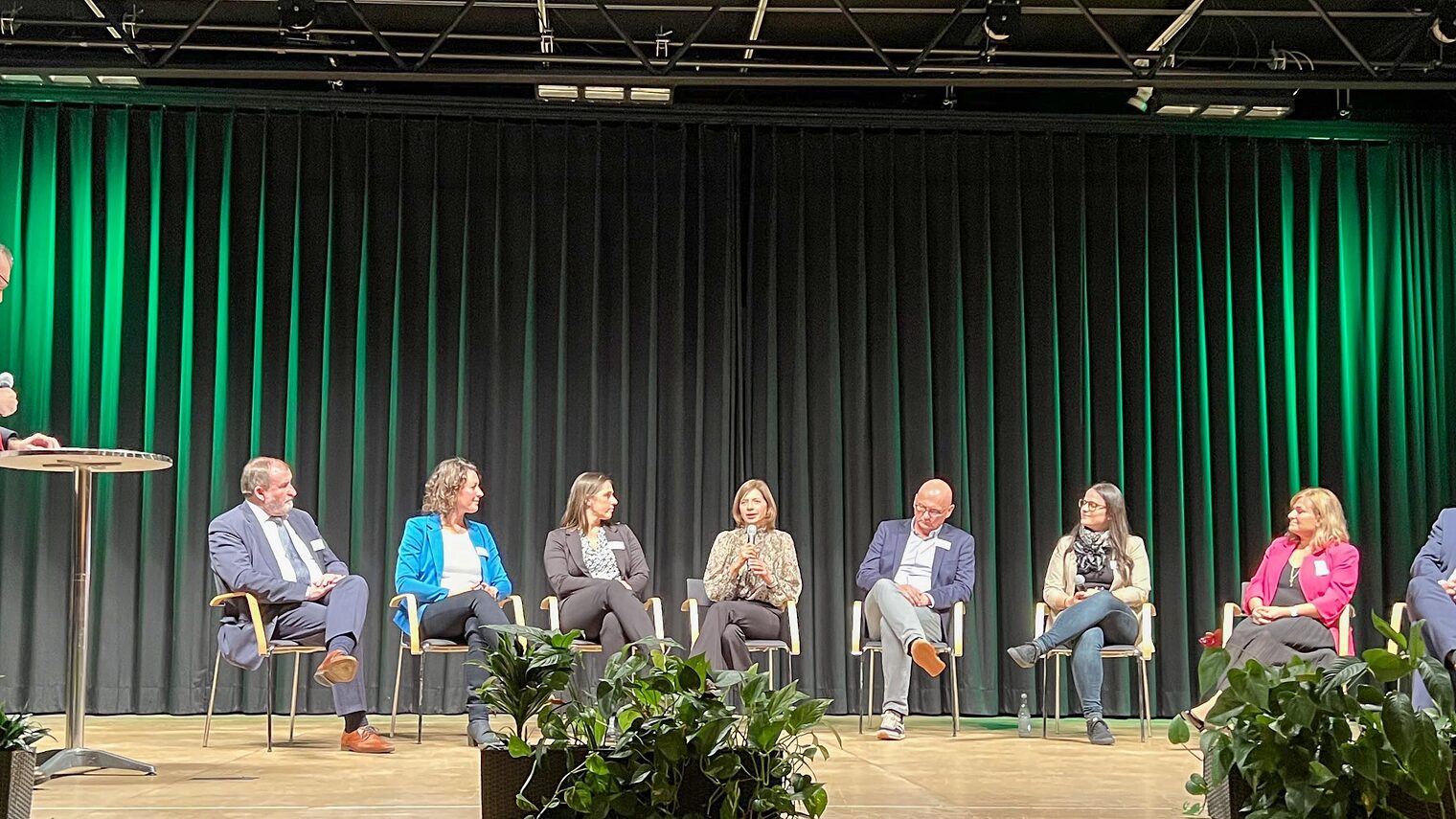 Personen sitzen in der Runde und diskutieren. Moderator steht am Pult mit Mikrofon in der Hand. 