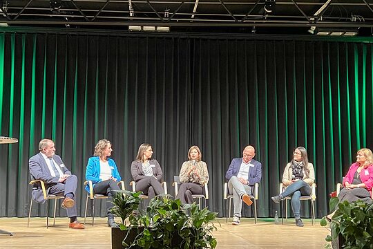 Personen sitzen in der Runde und diskutieren. Moderator steht am Pult mit Mikrofon in der Hand. 