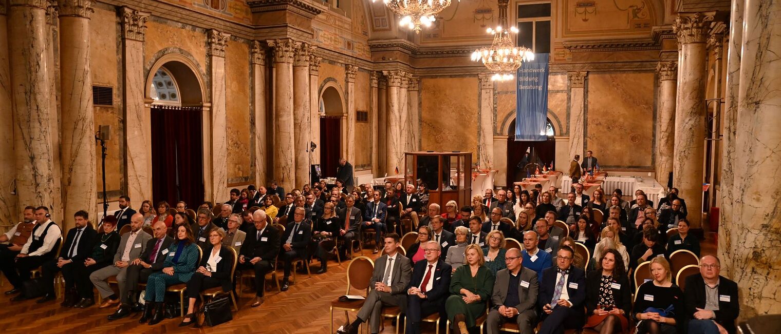 Menschen sitzen im großen, festlichen Saal und hören zu. 