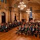 Menschen sitzen im großen, festlichen Saal und hören zu. 