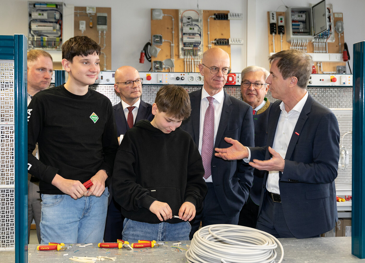 Männer und Jungen stehen zusammen in der Elektrowerkstatt. Zwei Jungen arbeiten an einem Kabel.