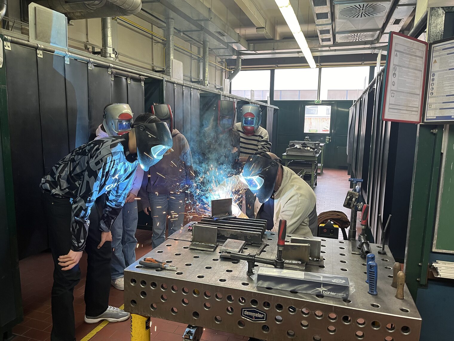 Schüler mit Schweißhelm übten direkt an einem Objekt in der Werkstatt. Blaue und rote Schweißfunken sprühen in die Luft. 