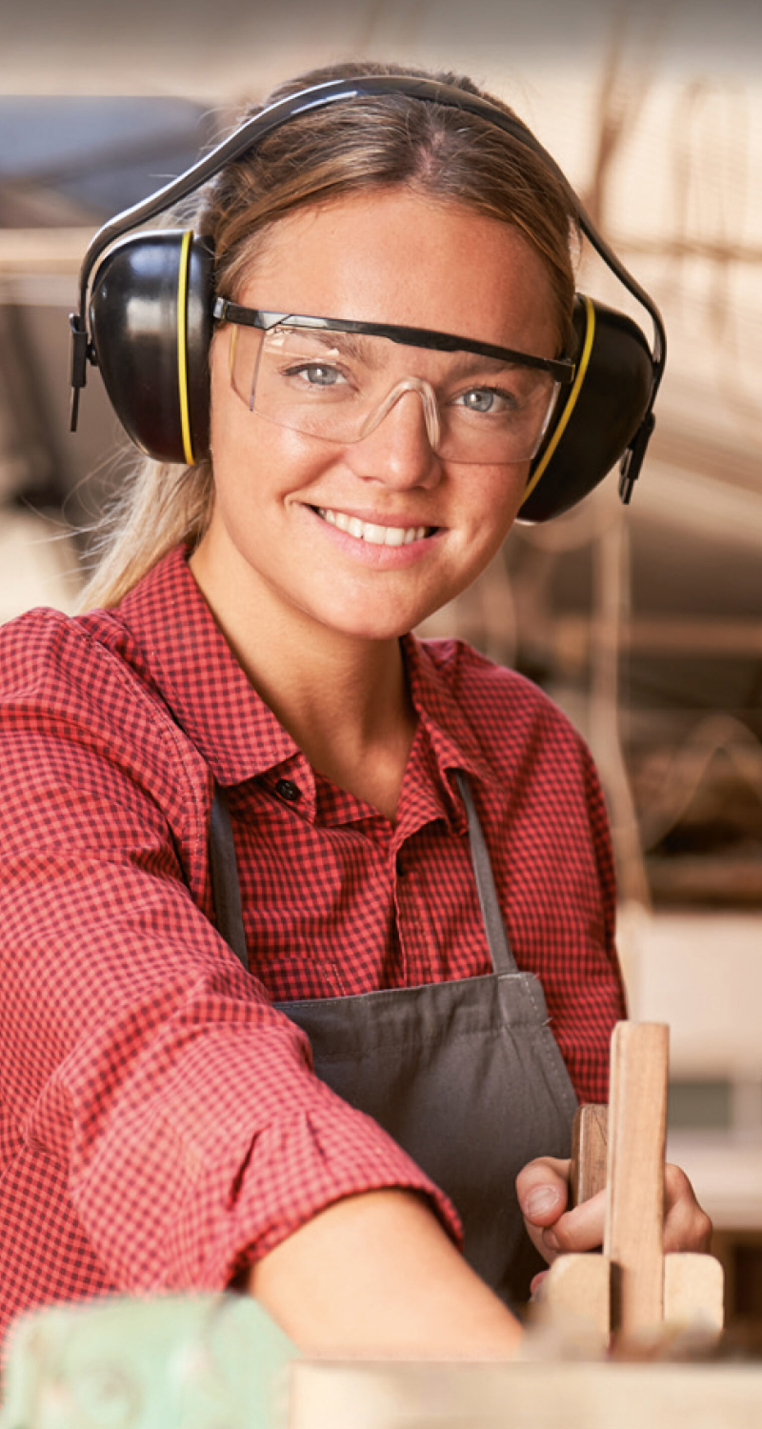 Eine lächelnde Schreinerin schleift Holz ab. Sie trägt eine Schutzbrille und Ohrenschützern.