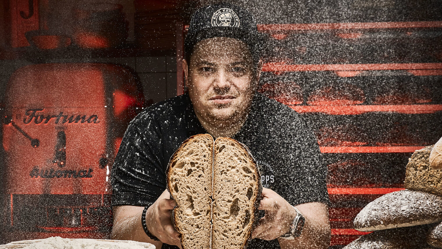 In einer rustikalen Bäckerei-Kulisse präsentiert Konditormeister Christian Philipps ein aufgeschnittenes Brot, während Mehlstaub in der Luft schwebt.