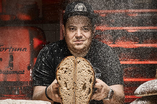 In einer rustikalen Bäckerei-Kulisse präsentiert Konditormeister Christian Philipps ein aufgeschnittenes Brot, während Mehlstaub in der Luft schwebt.