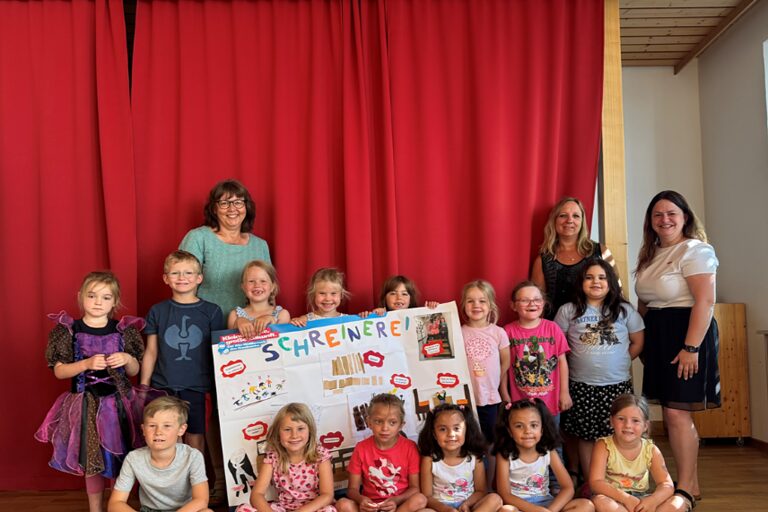Eine Kindergartengruppe mit zwei Erzieherinnen und der HWK-Vizepräsidentin Kathrin Zellner stehen vor einem roten Vorhang und halten ein Plakat zum Schreinerhandwerk.