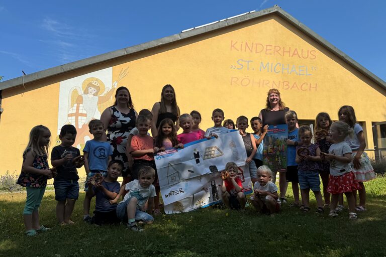 Die Kindergartenkinder, zwei Erzieherinnen und HWK-Vizepräsidentin Kathrin Zellner stehen vor dem gelben Kinderhaus St. Michael und halten ein großes Plakat.