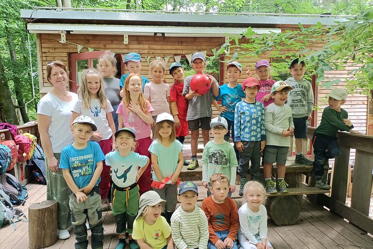 Die Kindergartenkinder stehen mit HWK-Vizepräsidentin Claudia Geiselhöringer auf einer Holzterasse vor einem Holzhäuschen.