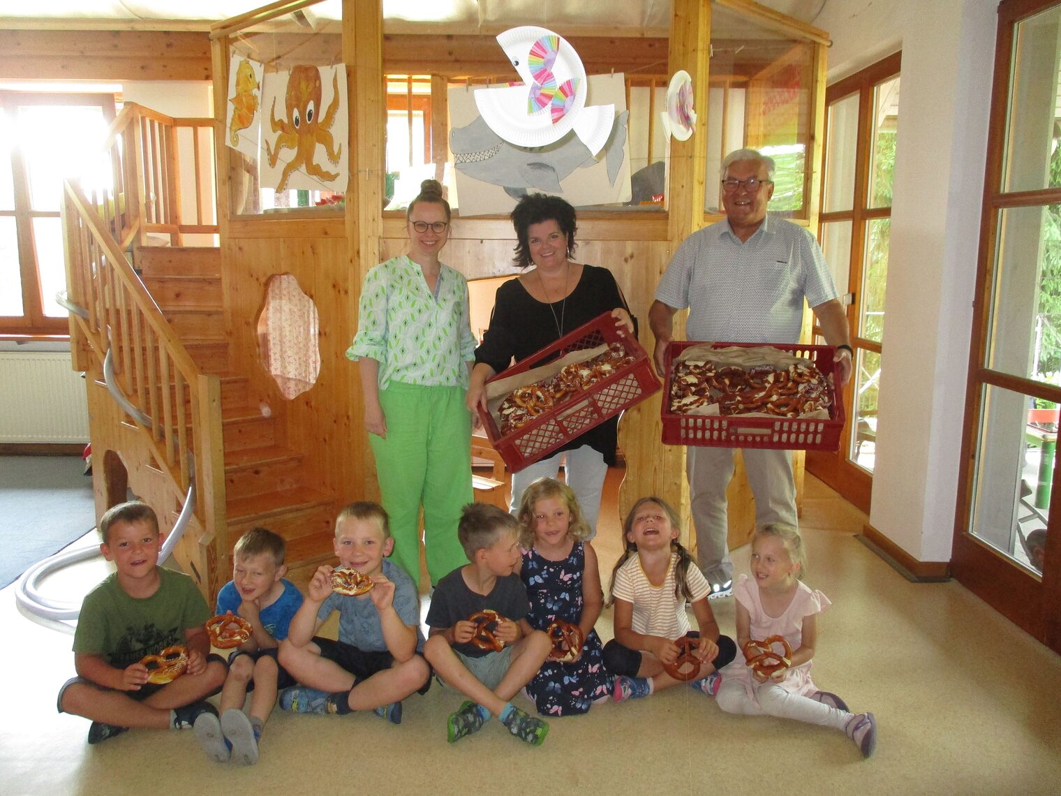 In einem hellen Raum stehen zwei Frauen und HWK-Vizepräsident Gerhard Ulm mit zwei Brezenkisten vor einem Holzspielhaus. Kinder sitzen am Boden und halten Brezen. 