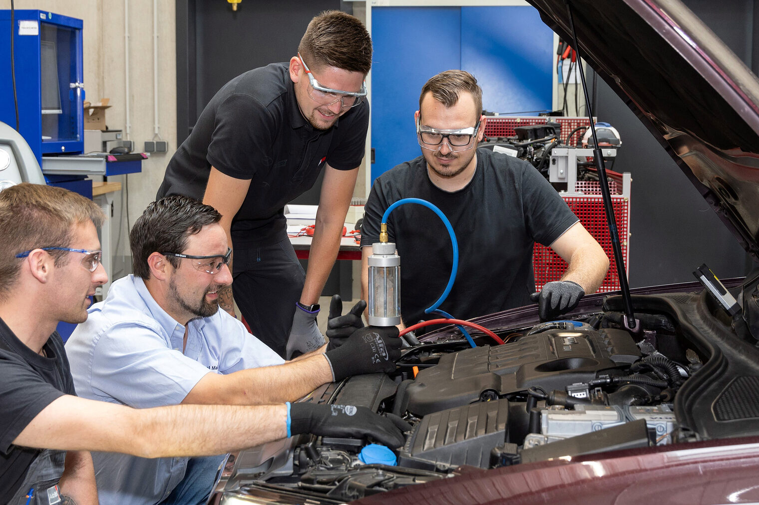 Drei Auszubildende im Kfz-Mechatronikerhandwerk untersuchen mit ihrem Ausbilder ein Auto in der KFZ-Werkstatt.