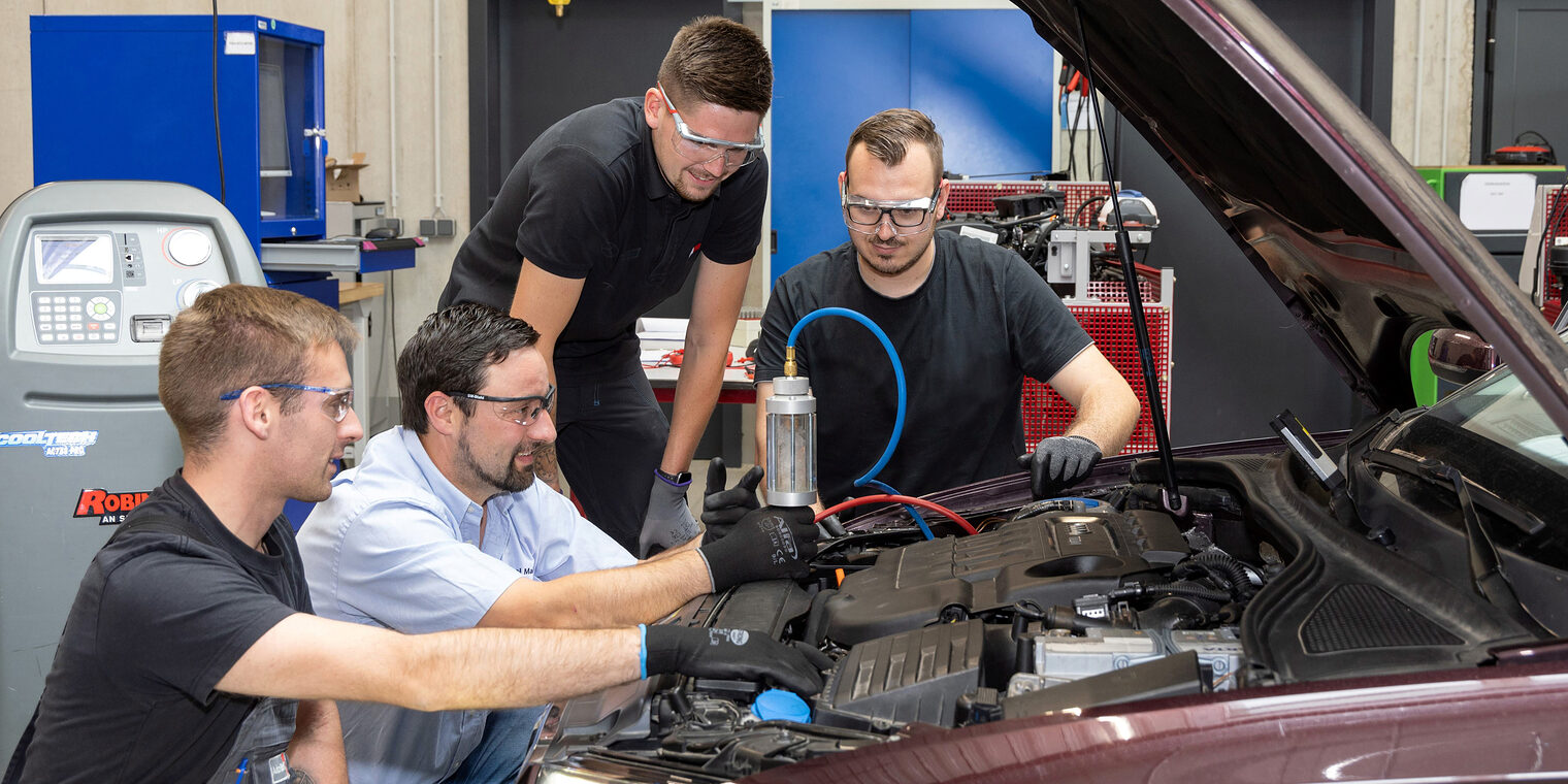Drei Auszubildende im Kfz-Mechatronikerhandwerk untersuchen mit ihrem Ausbilder ein Auto in der KFZ-Werkstatt.