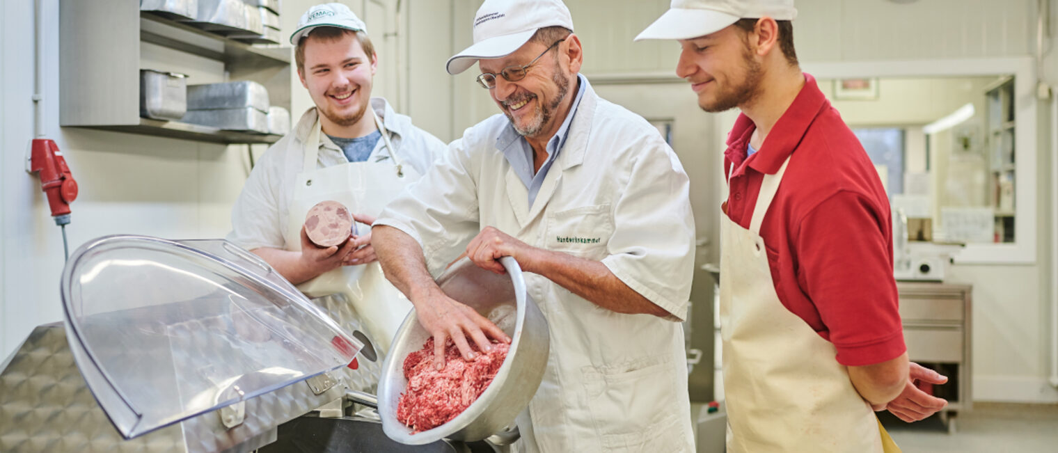 Unterweisung eines Ausbilders mit Auszubildenden an der Rühmaschine. Ein Ausbilder füllt Hackfleisch in einen Mischer, zwei Auszubildende sehen ihm dabei zu.