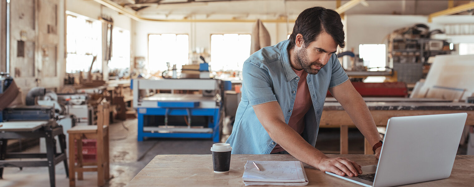 Der Betriebsinhaber steht in seiner Werkstatt vor dem Laptop und arbeitet.