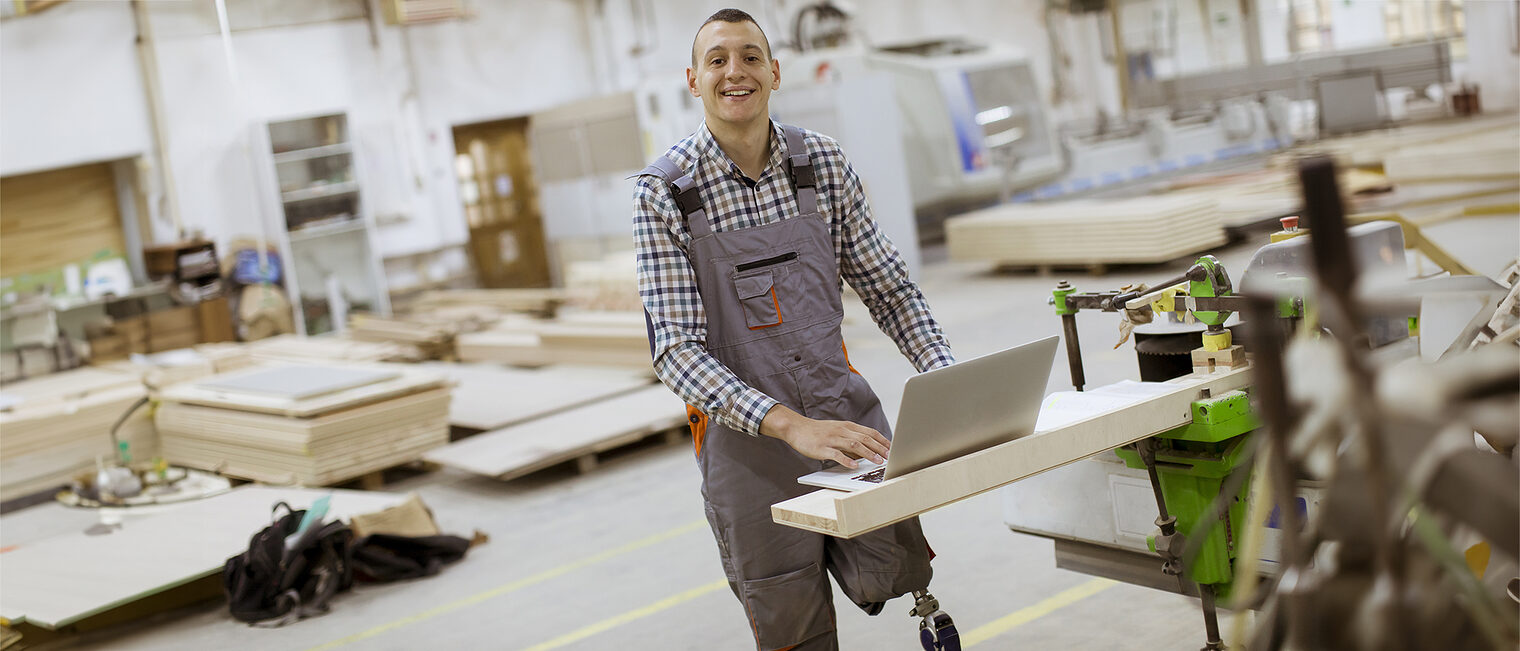 Ein Mann mit einer Beinprothese steht mit dem aufgeklappten Laptop in der Werkstatt. 
