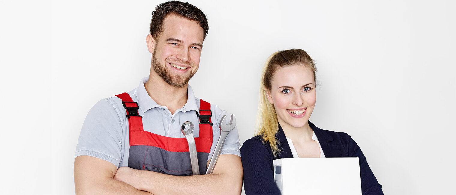 Ein junger Handwerker und eine junge Frau stehen selbstbewusst vor der Kamera. Er mit einem Schraubenschlüssel und sie mit einem Ordner. 