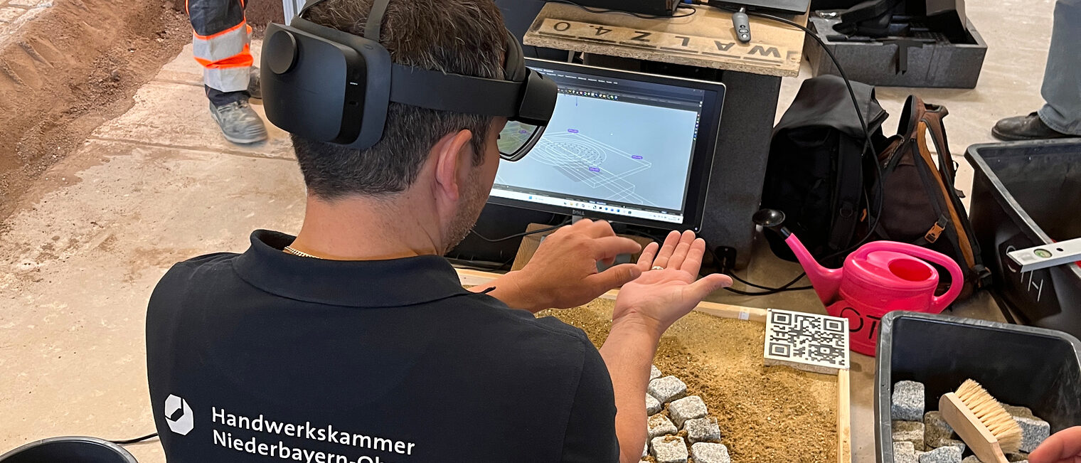 Ausbilder Ulrich Liebl hat eine VR-Brille aufgesetzt und kniet vor Pflastersteinen. Er übt damit virtuell pflastern.