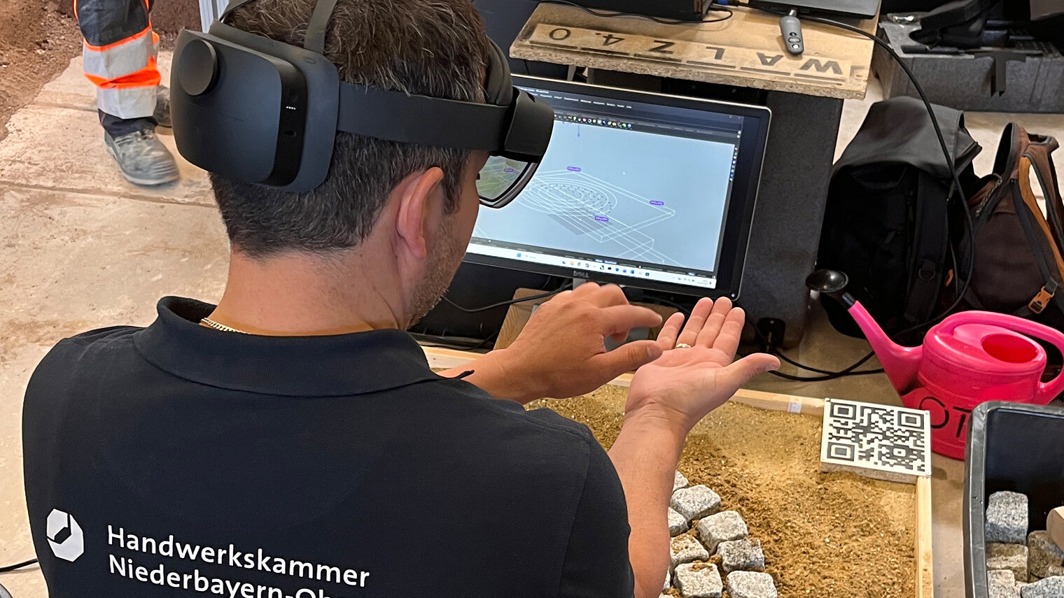 Ausbilder Ulrich Liebl hat eine VR-Brille aufgesetzt und kniet vor Pflastersteinen. Er übt damit virtuell pflastern.