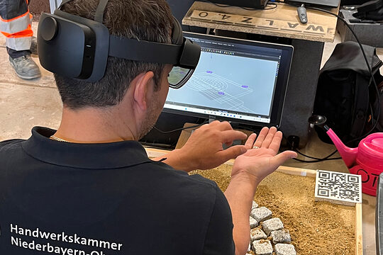 Ausbilder Ulrich Liebl hat eine VR-Brille aufgesetzt und kniet vor Pflastersteinen. Er übt damit virtuell pflastern.