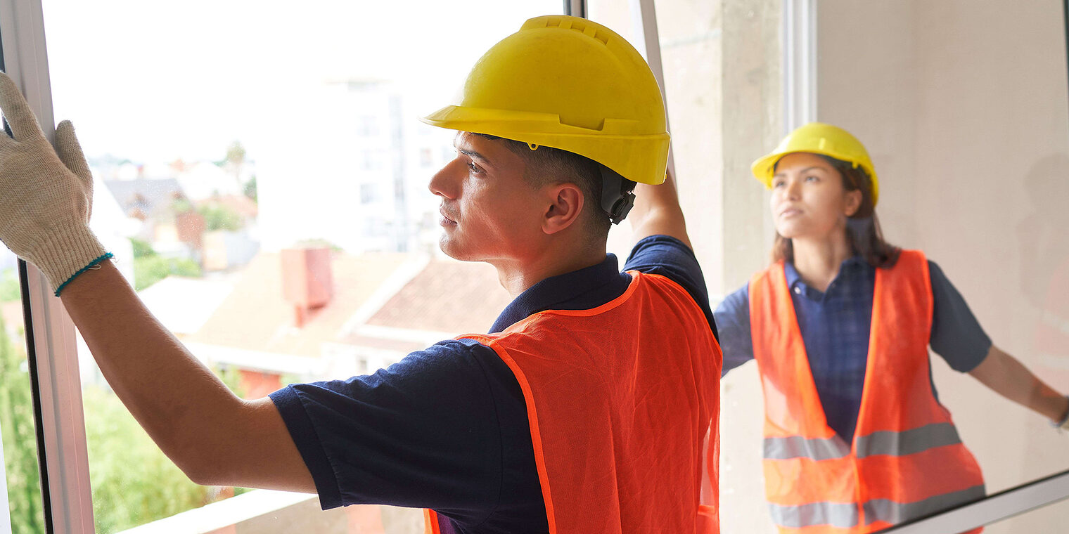 Ein Fensterbauer und eine Fensterbauerin montieren eine Balkontür in einem Neubau. Sie tragen Warnwesten und Helme.