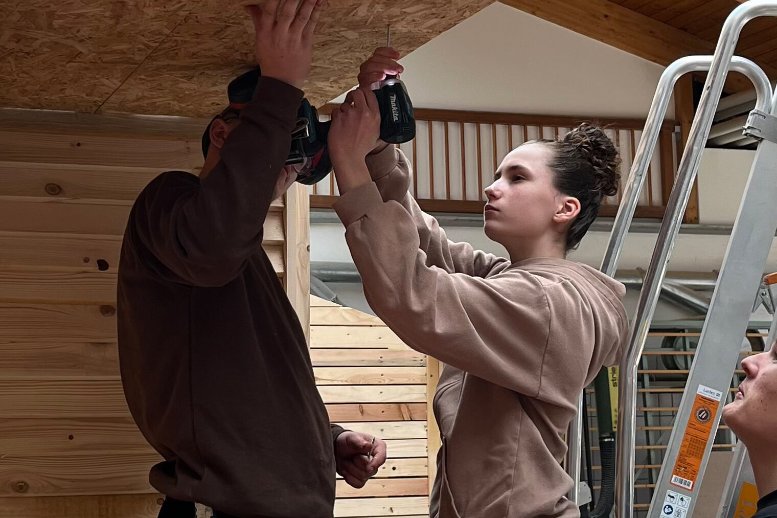 Ein Mädchen mit Bohrmaschine steht auf einer Leiter und arbeitet an einer Holzdecke; zwei Männer daneben geben Hilfestellung.
