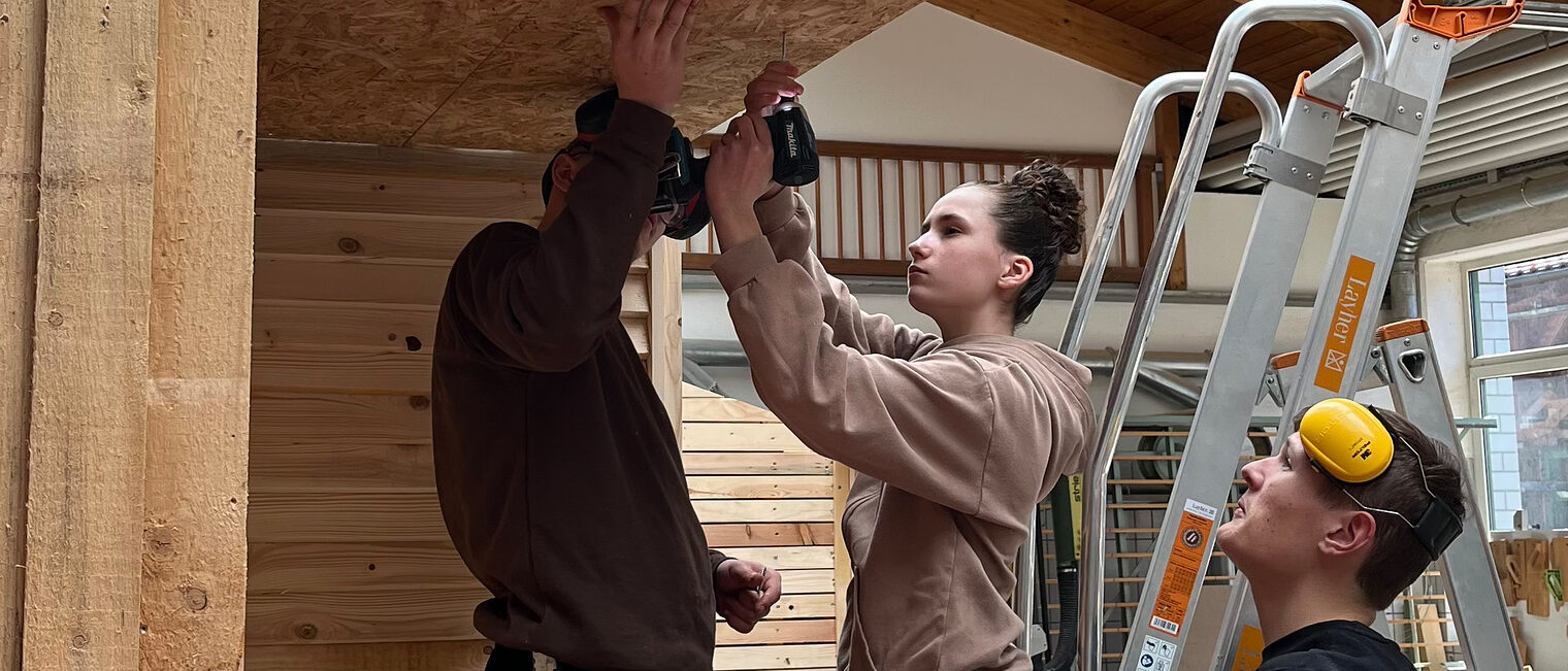Ein Mädchen mit Bohrmaschine steht auf einer Leiter und arbeitet an einer Holzdecke; zwei Männer daneben geben Hilfestellung.