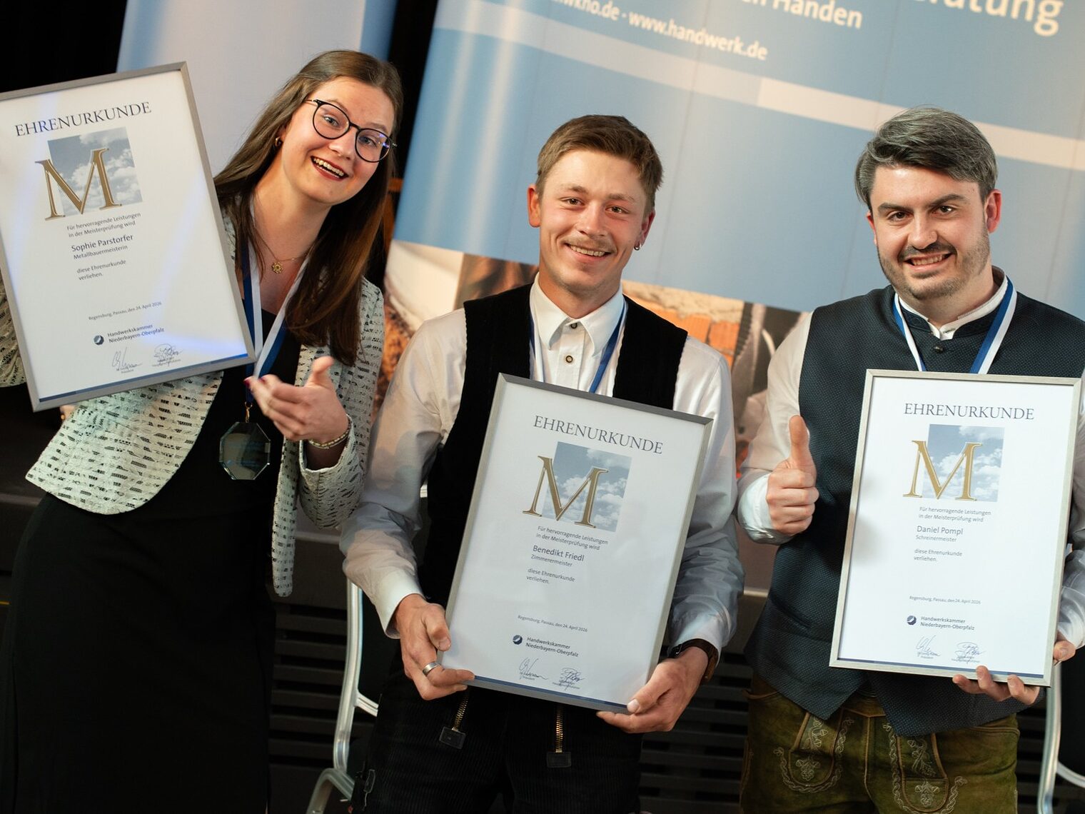 Metallbauermeisterin Sophie Parstorfer, Zimmerermeister Benedikt Friedl und Schreinermeister Daniel Pompl freuen sich über die Auszeichnung, strahlen in die Kamera und halten Urkunden. 