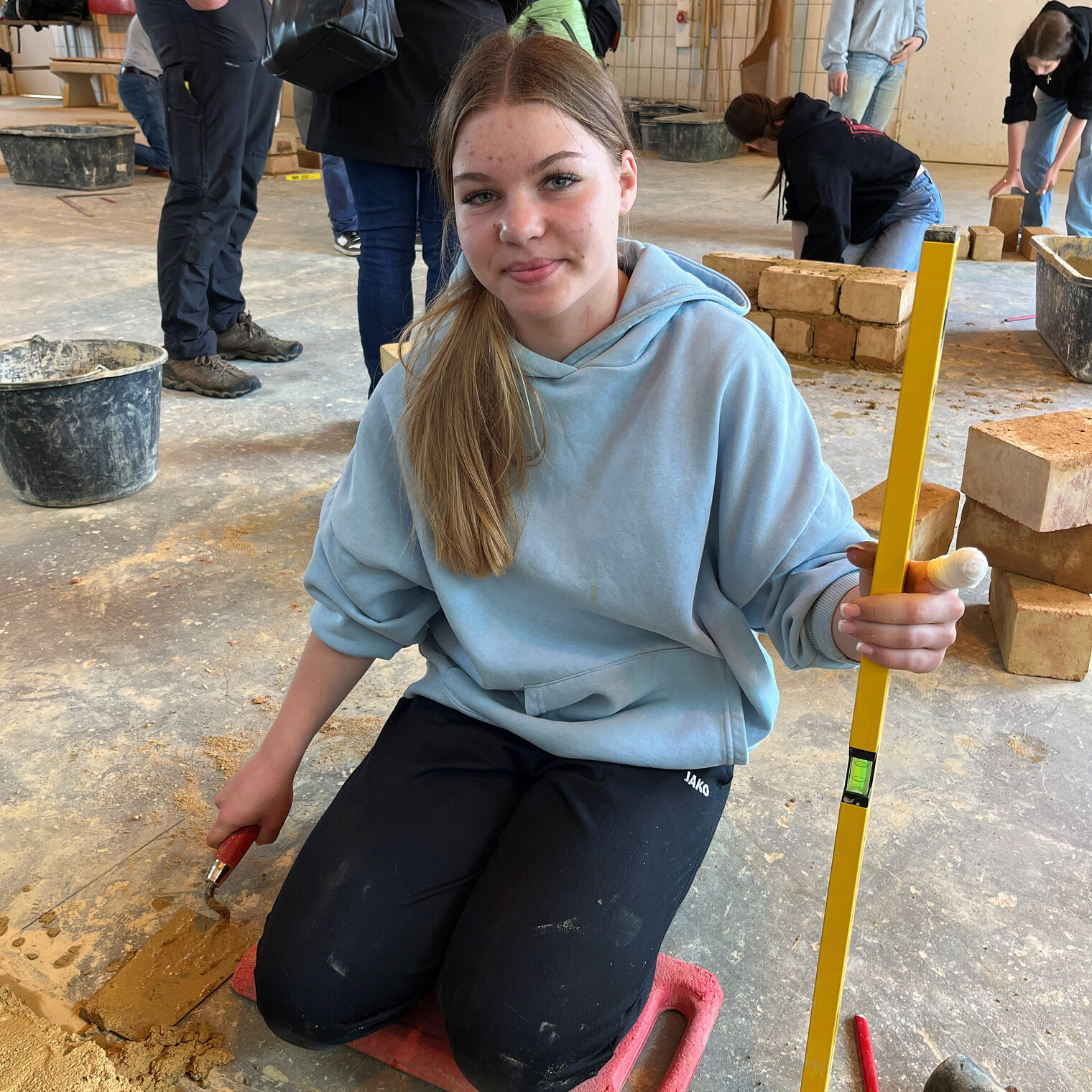 Hanna Neumaier zeigt ihr Können beim Mauern. Sie hält eine Wasserwaage in der linken Hand und mauert mit der rechten Hand. 