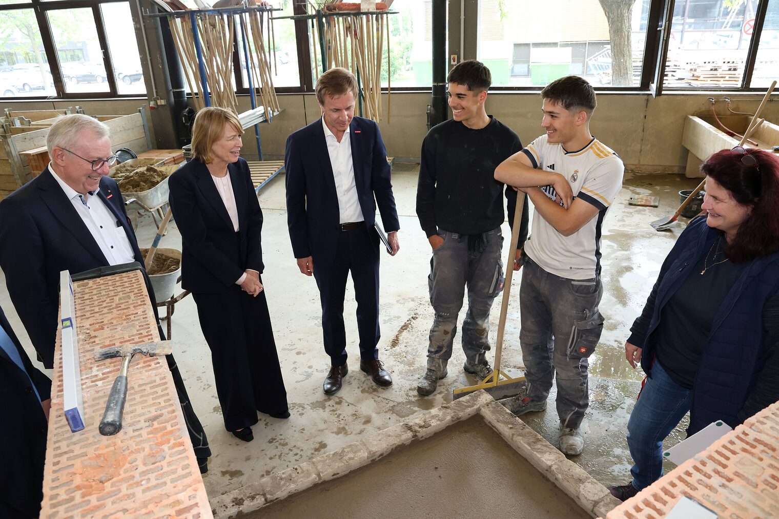 Elke Büdenbender im Gespräch mit zwei ÜLU-Teilnehmern im Maurer- und Betonbauerhandwerk, Ausbilderin Alexandra Stang, Jürgen Kilger und Hans Schmidt in der Werkstatt.